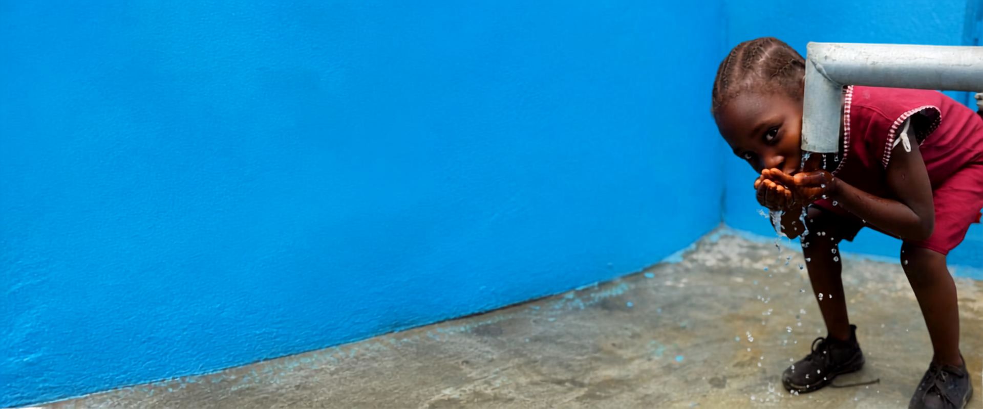 Kind drinkt schoon water met de handen bij een waterpomp tegen een blauwe muur