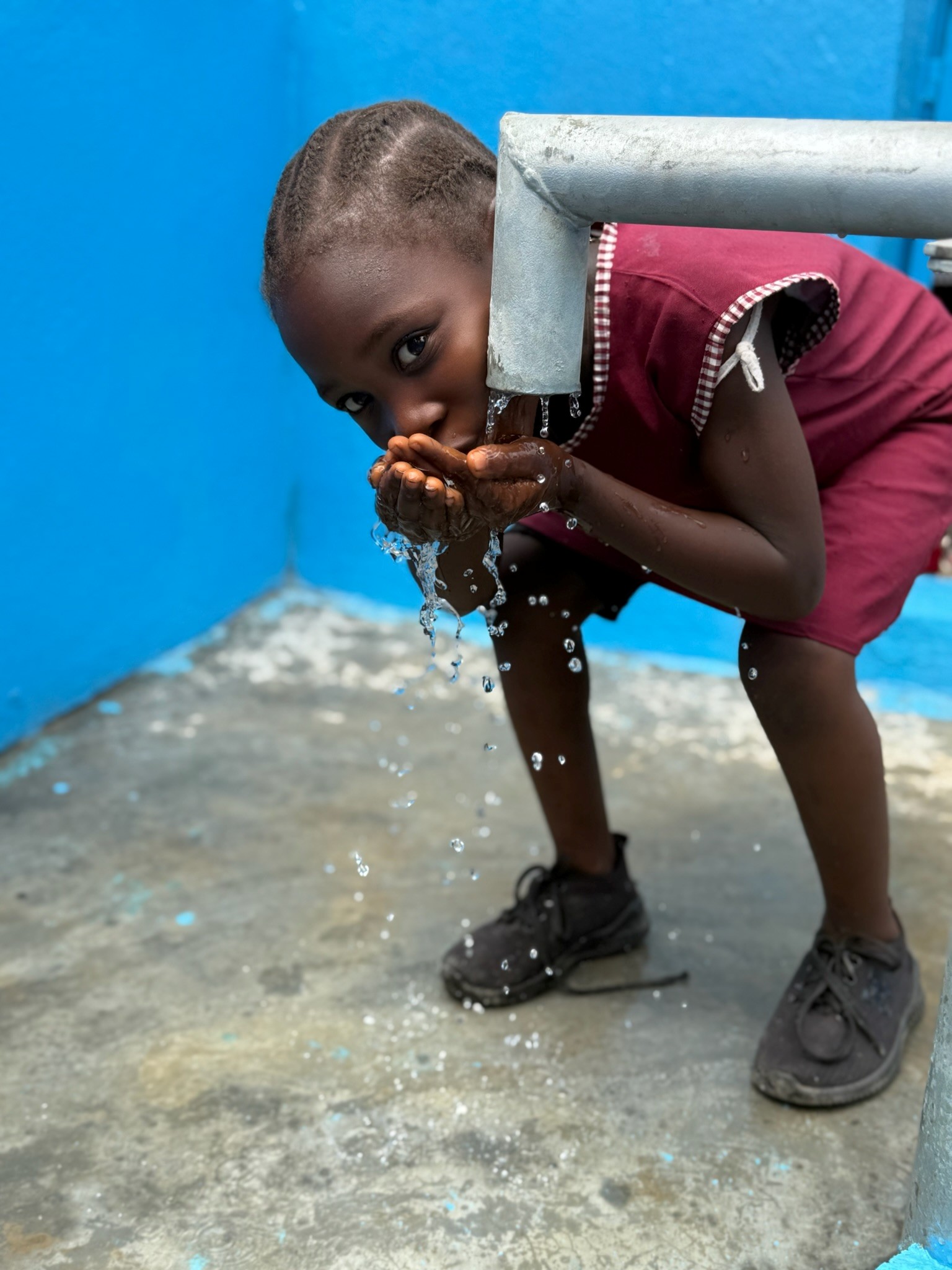 Kind drinkt schoon water met de handen bij een waterpomp tegen een blauwe muur